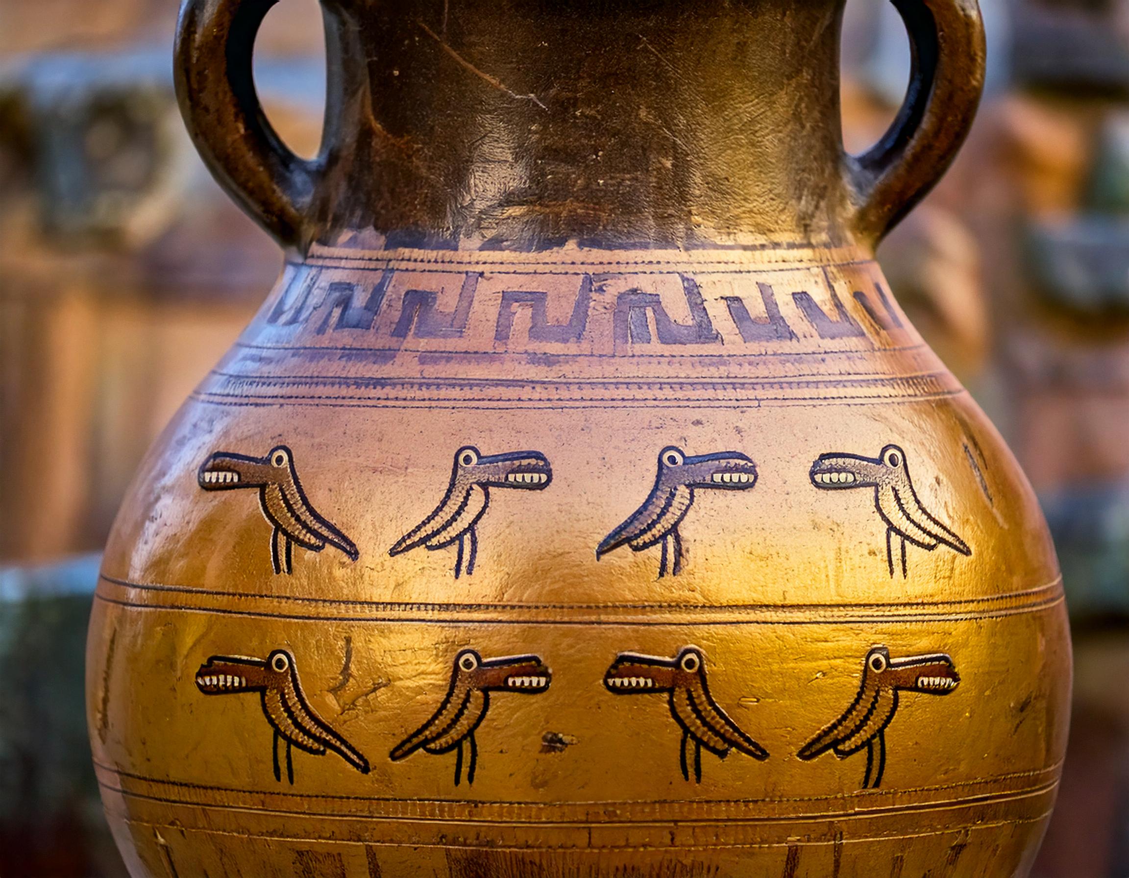 A copper vase with gold leaf and a dark lip matched by its shoulders. On it are Greek key patterns and many small Crocoducks.